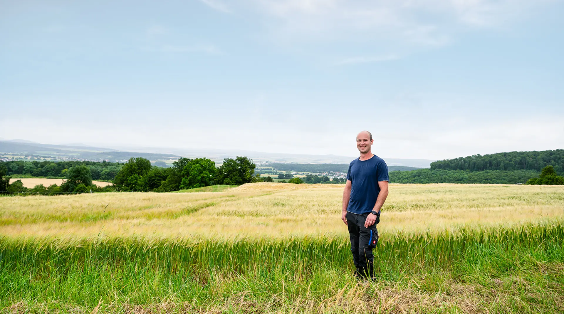 Junger Mann steht auf einer Wiese am Feldrand, im Hintergrund eine flache Landschaft mit Bäumen und ganz hinten am Horizont eine Ortschaft mit Häusern; darüber blauer Himmel mit leichten Wolken.