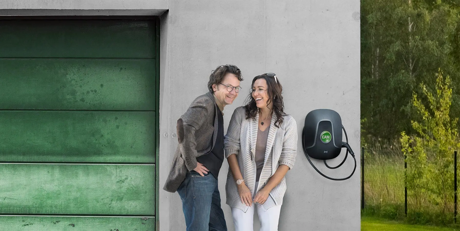 Ein Paar steht lächelnd vor einer grauen Wand mit einer grünen Garagentür und einer Elektroauto-Ladesäule daneben.
