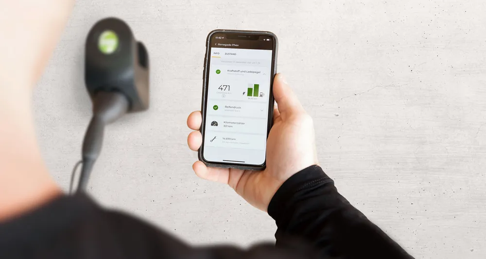 Hand hält Smartphone mit Anzeige zu Kilometerstand und Kraftstoffniveau, an einer grauen Wand.